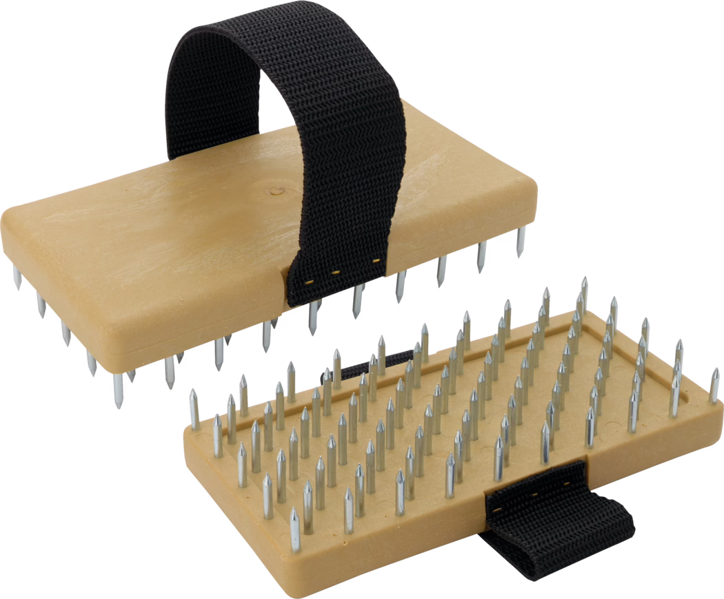 Pajarito_Dummy_Fine plaster scraper complet_033491_2021-03 Une vue détaillée d'un dispositif de massage pour les pieds comportant deux bases en bois plates avec des pointes métalliques. Le dispositif est doté d'une sangle en tissu noir robuste pour le fixer au pied, augmentant la stabilité pendant l'utilisation. La pièce supérieure est légèrement surélevée par rapport à la pièce inférieure, qui est entièrement couverte de pointes métalliques pour exercer une pression thérapeutique sur le pied. Le design favorise la relaxation et le soulagement de la douleur par l'acupressure.