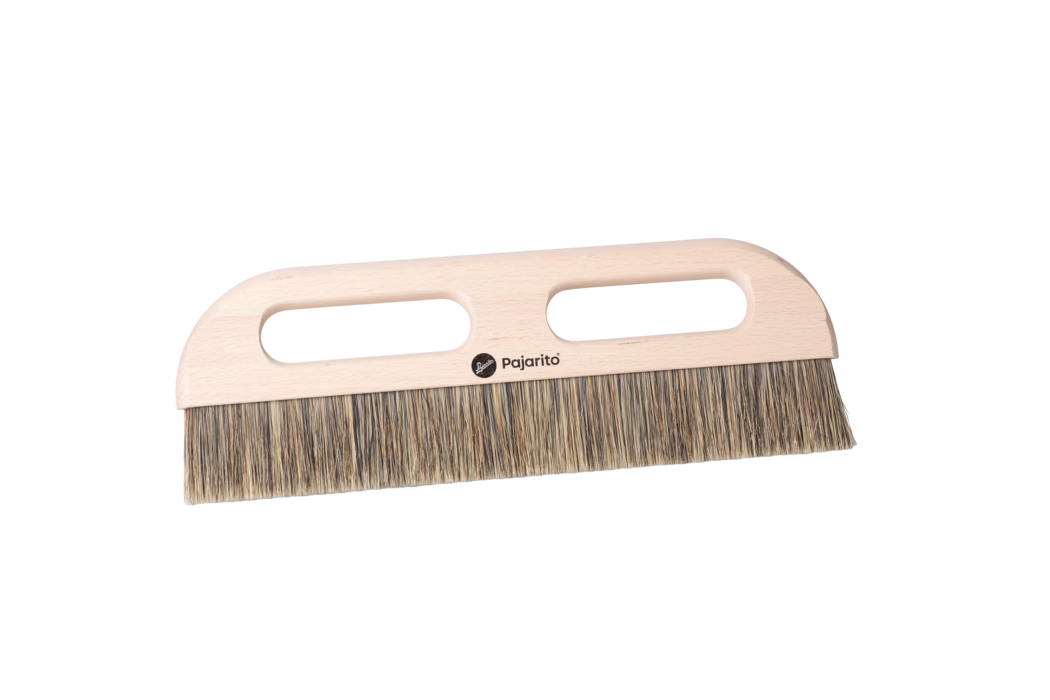 Une brosse en bois avec une tête plate et des poils, avec deux poignées découpées pour une meilleure prise. Les poils sont denses et légèrement variés en couleur, suggérant un matériau naturel, tandis que la base en bois semble lisse et polie avec un logo imprimé au centre.